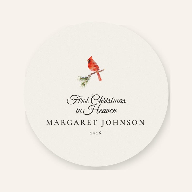 Front product card showing the complete round First Christmas in Heaven acrylic ornament on a warm cream background with a soft shadow. A watercolor red cardinal rests on a snow covered pine branch above the memorial wording, personalized name Margaret Johnson and year 2026. The uncluttered view makes the front artwork and circular edge easy to inspect.