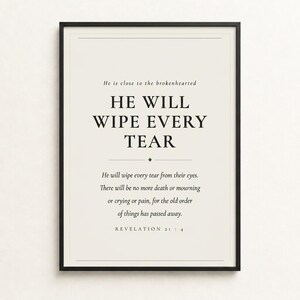 Styled shelf scene with the framed design beside an open Bible, natural botanicals, ceramics, and warm ambient light. The He Will Wipe Every Tear artwork presents Revelation 21:4 in minimalist black typography on warm beige. This Revelation 21:4 print suits a sympathy gift, memorial space, prayer room, bedroom, or comforting Christian home as printable digital wall art.
