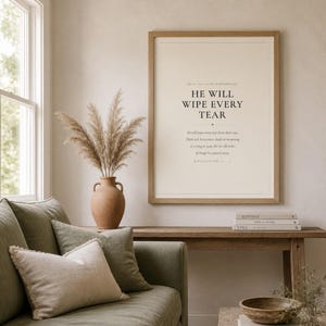 Warm lifestyle room with the framed design on a soft cream wall beside neutral decor and natural window light. The He Will Wipe Every Tear artwork presents Revelation 21:4 in minimalist black typography on warm beige. This Revelation 21:4 print suits a sympathy gift, memorial space, prayer room, bedroom, or comforting Christian home as printable digital wall art.