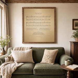 Amazing Grace vintage sheet music wall art in a rustic warm-oak frame, hanging above a sage-green velvet sofa with cream linen cushions and chunky knit throw blanket. Cozy farmhouse Christian living room with side table holding dried eucalyptus and an open hymnal Bible. Printable John Newton 1779 hymn from TheFramedVerse on Etsy.