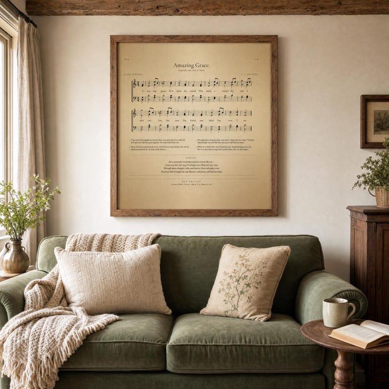 Amazing Grace vintage sheet music wall art in a rustic warm-oak frame, hanging above a sage-green velvet sofa with cream linen cushions and chunky knit throw blanket. Cozy farmhouse Christian living room with side table holding dried eucalyptus and an open hymnal Bible. Printable John Newton 1779 hymn from TheFramedVerse on Etsy.