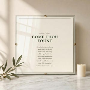 Come Thou Fount of Every Blessing hymn lyric print in floating glass frame with brass corner clips on marble surface with fresh olive branch and cream candle, dark forest green on warm cream, premium christian hymn wall art, come thou fount art song lyric art baptism confirmation first communion gift