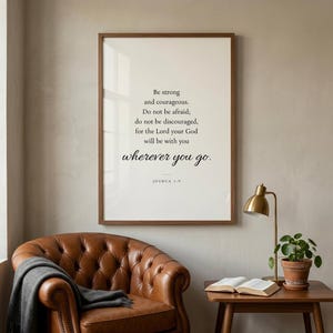 Joshua 1:9 'Be Strong and Courageous' Christian wall art in modern minimalist serif typography on cream background, framed in warm walnut above a cognac leather chair in a masculine Christian study with open leather Bible and brass lamp. Premium Father's Day digital download, gift for Christian dads, granddads, confirmation, graduation.
