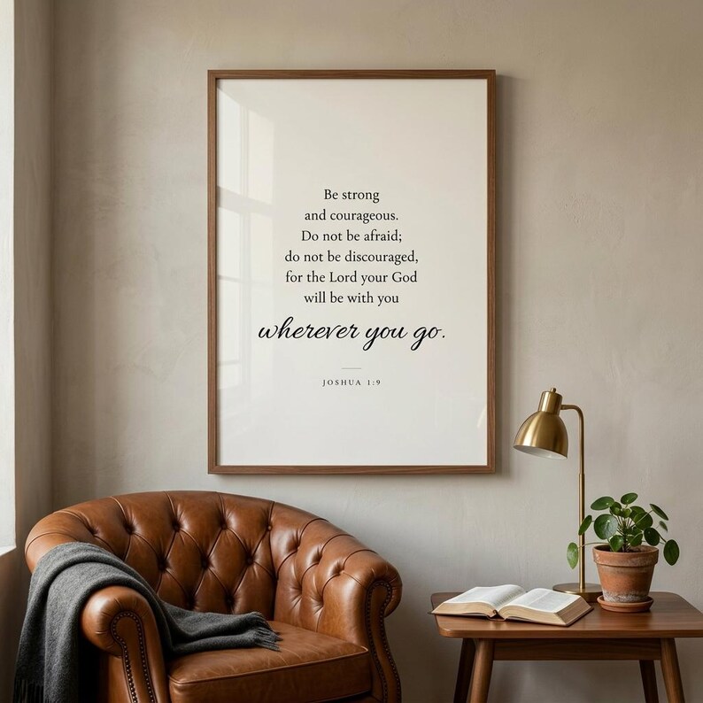 Joshua 1:9 'Be Strong and Courageous' Christian wall art in modern minimalist serif typography on cream background, framed in warm walnut above a cognac leather chair in a masculine Christian study with open leather Bible and brass lamp. Premium Father's Day digital download, gift for Christian dads, granddads, confirmation, graduation.