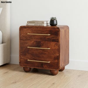 May include: A three-drawer wooden nightstand with a warm brown finish and brass-colored handles. The nightstand has rounded edges and sits on three small, rounded feet. A small potted succulent and two books are on top.