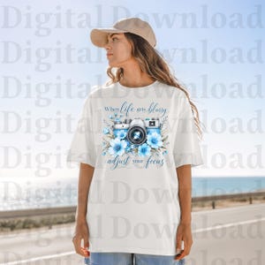 May include: White t-shirt featuring a blue and white camera design with floral accents. The text "When life gets blurry, adjust your focus" is printed above the camera. The shirt is a casual, everyday wear item.