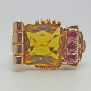 May include: A gold ring featuring a large, rectangular, faceted yellow citrine gemstone as the centerpiece. To the right of the citrine, three small, round, deep pink rubies are set vertically in a row. The metal band has a polished finish.