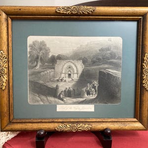 May include: A framed black and white print depicting an architectural scene with figures. The print is set against a teal background within a wooden frame with ornate gold-colored corner accents. The frame is approximately 16 inches wide.
