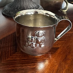 May include: A silver-colored baby cup with a handle and two embossed teddy bears on the side. The cup is sitting on a dark wooden surface, with other decorative items in the background.