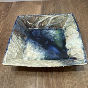 May include: A square ceramic serving dish with a unique, textured glaze. The dish features a blend of blue, white, and green hues, creating a marbled effect. The edges are slightly raised, and the overall design is modern and artistic.