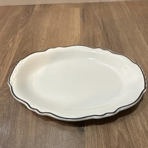 Puede incluir: Plato ovalado blanco con borde ondulado y ribete negro. El plato es de cerámica y se encuentra sobre una superficie de madera. Este plato es adecuado para servir aperitivos, entrantes o postres.