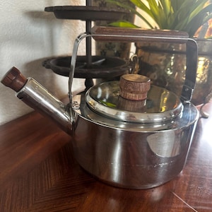May include: A stainless steel tea kettle with a wooden handle and lid knob. The kettle has a spout and a curved handle. The lid is circular and fits snugly on top. The wooden handle and knob provide a contrast to the shiny metal.
