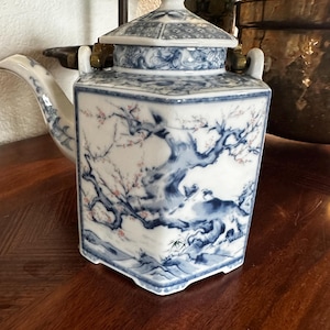Könnte beinhalten: Eine weiß-blaue Porzellan-Teekanne mit quadratischem Korpus und Deckel. Die Teekanne hat ein detailliertes Landschaftsdesign mit Bäumen und Blumen. Ausguss und Henkel sind gebogen, und der Deckel hat einen kleinen Knopf.