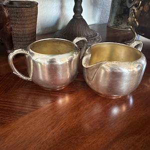 May include: Two silver-coloured serving pieces, a creamer and a sugar bowl, sit on a wooden surface. The creamer has a spout and handle, while the sugar bowl has a handle. Both have a gold-coloured interior.