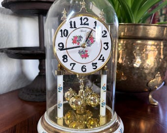 Op de afbeelding: Een vintage klok met een witte wijzerplaat, zwarte cijfers en bloemaccenten, ingekapseld in een glazen koepel. De klok heeft een goudkleurig frame en slinger en staat op een witte basis met gouden rand. Het is een decoratief woondecoratie-item.