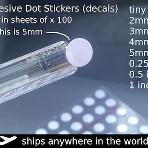 May include: A close-up of a white adhesive dot sticker, held by a clear plastic tool. The image includes text indicating the stickers are sold in sheets of 100 and are available in various sizes, including 0.25, 0.5, and 1 inch.