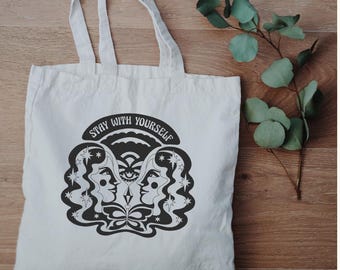 May include: A white canvas tote bag featuring a black graphic print of two faces in profile, stars, a butterfly, and an eye symbol. The text "STAY WITH YOURSELF" is printed in a curved arch above the central illustration on the front of the bag.