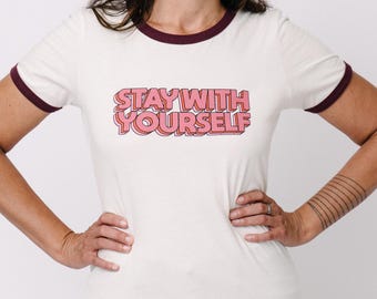 May include: A cream-colored ringer t-shirt with dark maroon trim on the collar and sleeve cuffs. The center of the shirt features the text "STAY WITH YOURSELF" in a bold, pink, three-dimensional block font with a dark outline.