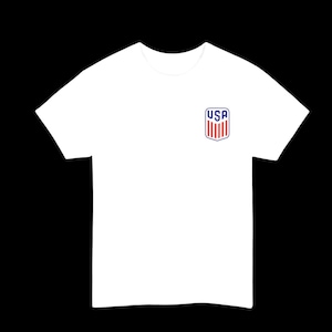 Puede incluir: Una camiseta blanca con un pequeño emblema de USA en el pecho. El emblema es un diseño de escudo con rayas rojas y blancas y la palabra "USA" en azul.