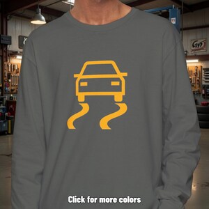 Pode incluir: Uma camisola cinzento-carvão de manga comprida com um gráfico amarelo centrado de um carro com duas linhas onduladas por baixo, representando um símbolo de aviso de controlo de tração. O texto "Click for more colors" está na parte inferior.