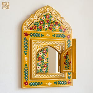 Puede incluir: Un espejo decorativo amarillo pintado a mano con una parte superior arqueada y una pequeña puerta. El espejo está adornado con patrones florales en rojo, blanco y verde. El marco presenta intrincados detalles blancos y el texto "MARCU DECOR".
