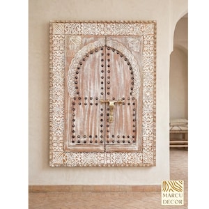 Puede incluir: Panel decorativo de pared de madera con acabado envejecido. El panel presenta un diseño de puerta arqueada con tachuelas metálicas y un pestillo de latón. Intrincadas tallas blancas enmarcan las puertas, agregando elegancia. El panel es de un color cálido y neutro.