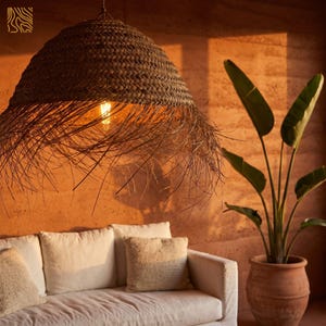 May include: A woven, brown pendant light fixture hangs above a white sofa with beige pillows. The light fixture has a fringed edge. A potted plant with large green leaves sits to the right of the sofa. The wall is a warm orange color.