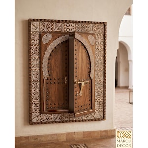 Puede incluir: Una puerta de madera montada en la pared con una parte superior arqueada y intrincados diseños blancos tallados. La puerta está abierta, revelando un vistazo del interior. El marco presenta patrones geométricos detallados. Un pestillo de latón y tachuelas decorativas se suman al diseño.