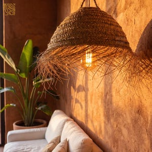 May include: A woven straw pendant light fixture with a fringed edge hangs from the ceiling. The light bulb is visible inside the shade, casting a warm glow. The fixture is suspended above a white sofa and a potted plant.