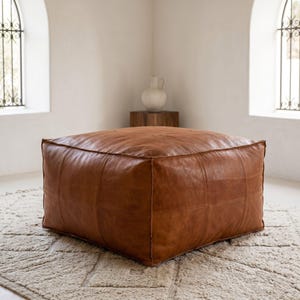 Könnte beinhalten: Ein großer, quadratischer Leder-Sitzhocker in Cognac mit sichtbaren Nähten auf der Oberfläche. Das Möbelstück besteht aus mehreren Lederbahnen, die eine strukturierte, flache Würfelform mit einer glatten, matten Oberfläche bilden.