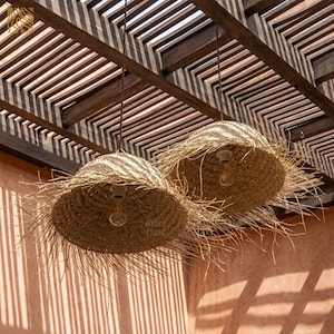 May include: Two pendant lights with woven straw shades hang from a wooden pergola. The shades have a fringed edge and house exposed light bulbs. The background is a peach-colored wall with shadows.