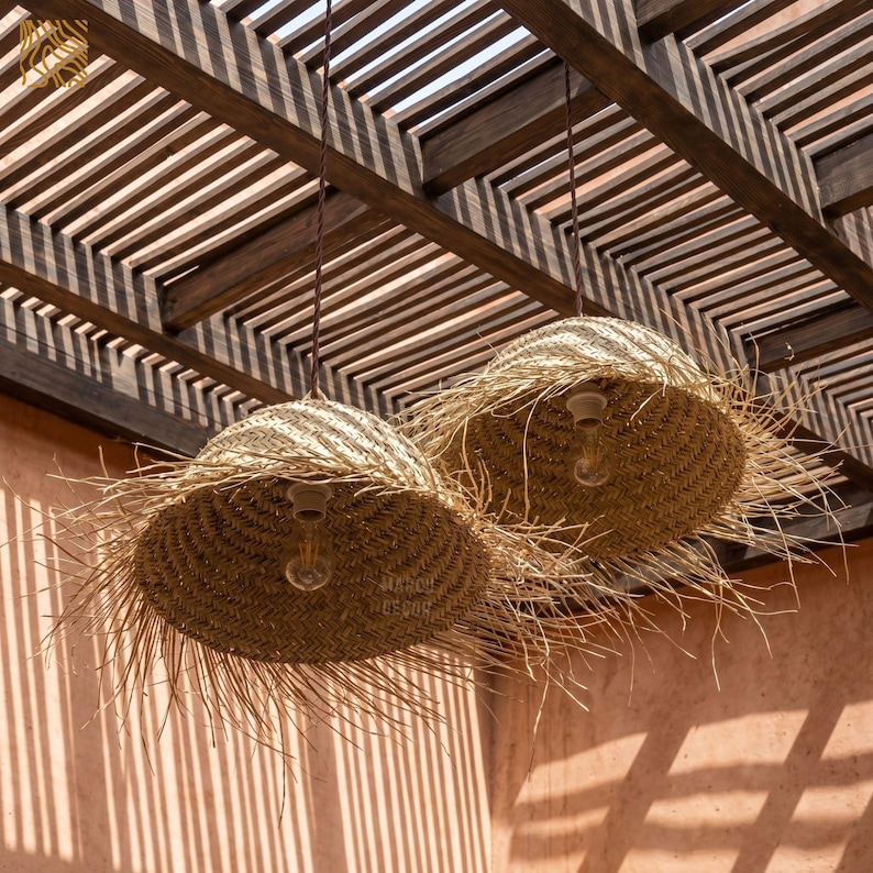 May include: Two pendant lights with woven straw shades hang from a wooden pergola. The shades have a fringed edge and house exposed light bulbs. The background is a peach-colored wall with shadows.