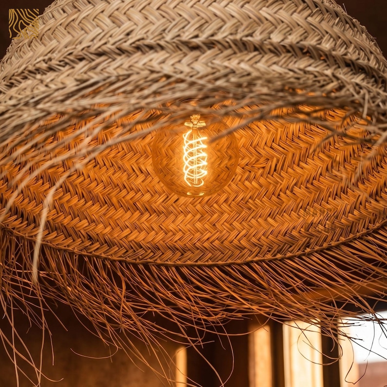 May include: Close-up of a woven straw lampshade with a warm, glowing lightbulb. The lampshade has a natural, textured appearance with a fringed edge. The lightbulb is a vintage style with a spiral filament.