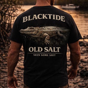 Könnte beinhalten: Schwarzes T-Shirt mit einer detaillierten Illustration eines Alligators und dem Text "BLACKTIDE OLD SALT SEEN SOME SHIT" in einer Used-Schriftart. Das T-Shirt wird von einer Person im Freien getragen.