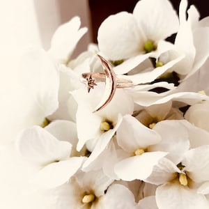 Op de afbeelding: Een ring van goudkleurig metaal met een halve maan ontwerp en een kleine ster aan de bovenkant. De ring ligt op een bed van witte bloemen.