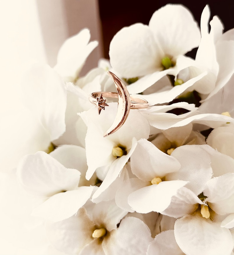 Op de afbeelding: Een ring van goudkleurig metaal met een halve maan ontwerp en een kleine ster aan de bovenkant. De ring ligt op een bed van witte bloemen.