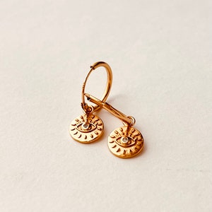 May include: Gold hoop earrings with a dangling charm featuring a gold eye with a small diamond in the center.