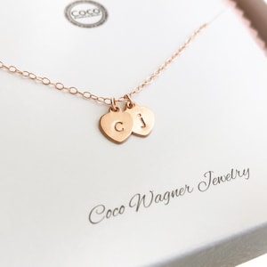 May include: A rose gold necklace with two small heart charms, each with a letter engraved on it. The letters are "C" and "J". The necklace is in a white box with the words "Coco Wagner Jewelry" printed on it.