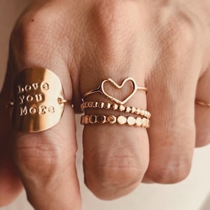 May include: A set of three gold rings. The first ring is a wide band with the words "Love You More" engraved on it. The second ring is a delicate open heart design. The third ring is a band of small, round beads.