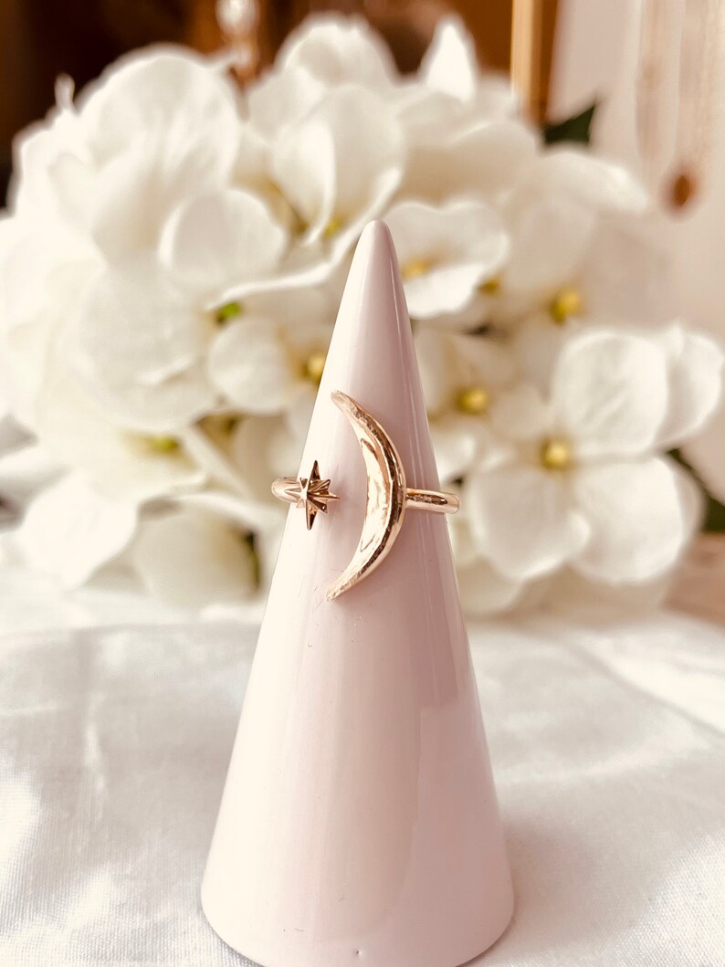 Op de afbeelding: Een gouden halve maan ring met een klein sterretje detail. De ring wordt getoond op een witte, kegelvormige ringhouder.