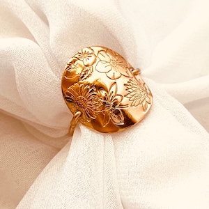 May include: A gold-colored ring with a large, oval-shaped face. The ring's face is intricately decorated with embossed floral designs, including sunflowers and other detailed blossoms. The ring is set against a backdrop of soft, white fabric.