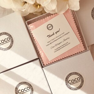 Op de afbeelding: Drie witte cadeaudozen met een roze poetsdoek in een van de dozen. De dozen hebben een zwart-wit logo met de tekst "Coco Wagner Jewelry". De poetsdoek heeft hetzelfde logo en de tekst "Bedankt! Draag het. Deel het. Vertel ons van je liefde! Volg @CocoWagner Jewelry op Instagram Coco Wagner Jewelry CocoWagnerJewelry.com CocoWagner.Jewelry.Etsy.com"