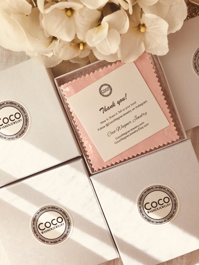 Op de afbeelding: Drie witte cadeaudozen met een roze poetsdoek in een van de dozen. De dozen hebben een zwart-wit logo met de tekst "Coco Wagner Jewelry". De poetsdoek heeft hetzelfde logo en de tekst "Bedankt! Draag het. Deel het. Vertel ons van je liefde! Volg @CocoWagner Jewelry op Instagram Coco Wagner Jewelry CocoWagnerJewelry.com CocoWagner.Jewelry.Etsy.com"