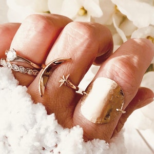 Op de afbeelding: Een hand met een set gouden ringen, waaronder een halvemaanvormige ring, een stervormige ring en een grote, ronde, gehamerde ring.