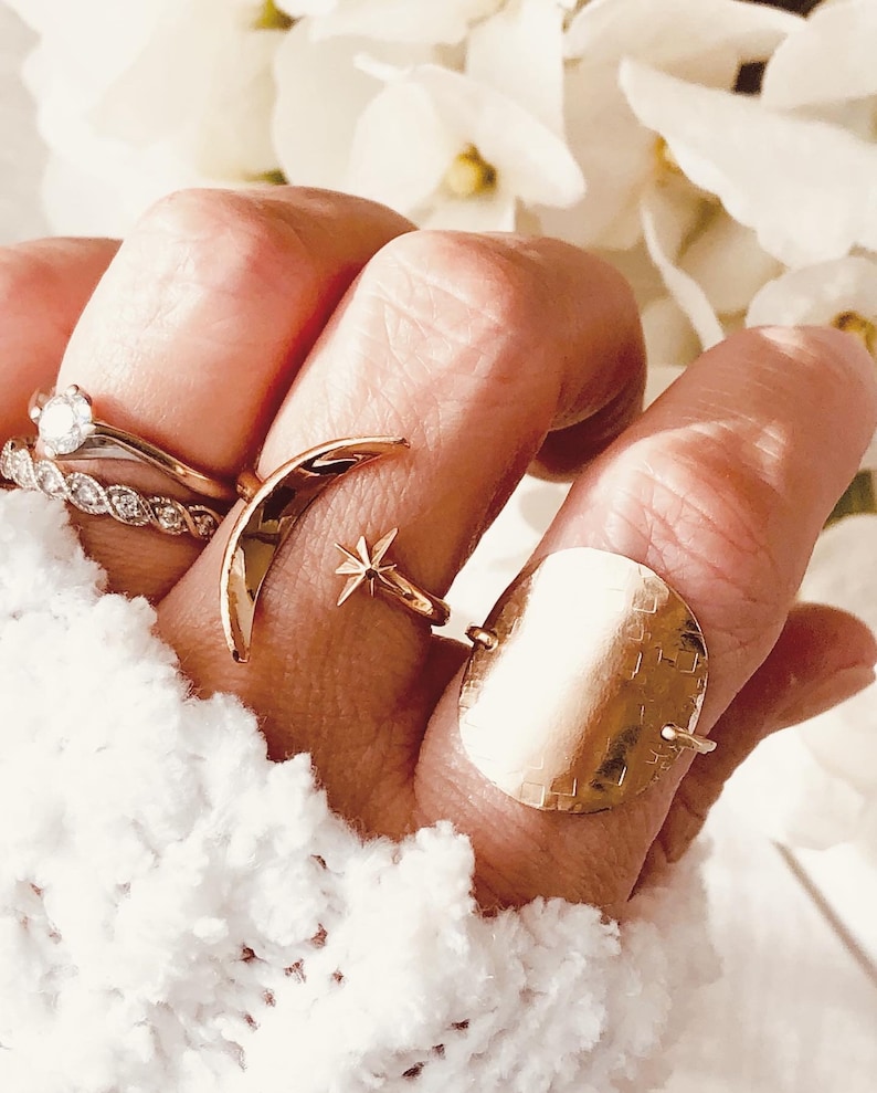 Op de afbeelding: Een hand met een set gouden ringen, waaronder een halvemaanvormige ring, een stervormige ring en een grote, ronde, gehamerde ring.