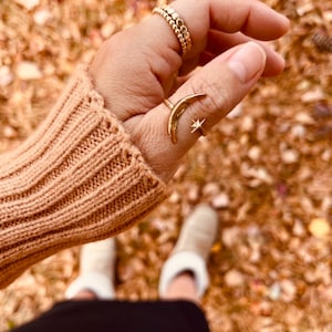 Op de afbeelding: Een hand draagt een gouden halvemaanring met een ster op de duim en een gouden kralenring op de wijsvinger. De hand draagt een bruine trui en de persoon draagt witte schoenen.