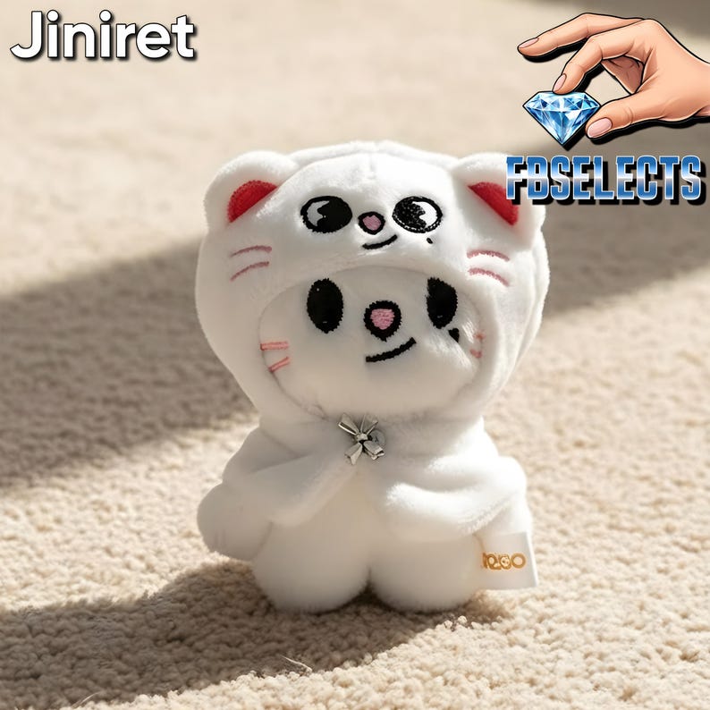 May include: A white plush toy in a cat costume with red accents on the ears and pink whiskers. The toy has black eyes and a small silver bow. The text "JINIRE" is at the top left and "FBSELECTS" is at the top right.