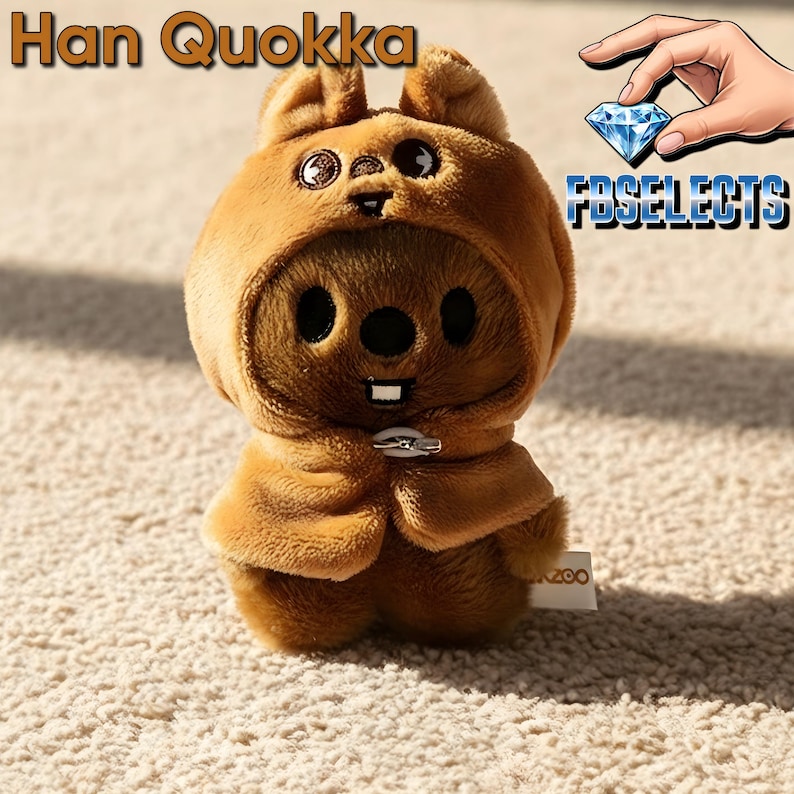 May include: A plush brown Quokka toy with a hooded outfit and a small cape, secured with a silver clasp. The toy has large black eyes, a round nose, and a single white tooth. The text "Han Quokka" is at the top, and "FBSELECTS" is in the background.