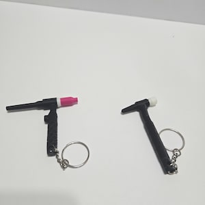 May include: Two black and silver TIG welding torch keyrings. One has a pink tip, the other a white tip. Each keyring has a silver key ring attached to the handle.