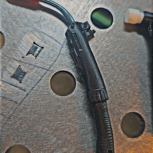 May include: A black welding torch with an orange tip and a silver metal neck. The torch is attached to a metal surface with several circular holes. A silver keyring is attached to the bottom of the torch.
